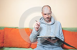 Young bald man in casual clothes with funny and confused facial expression trying to calculate his year tax debt with pen and pape