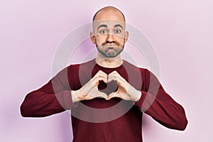 Young bald man doing heart symbol with hands puffing cheeks with funny face