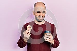 Young bald man drinking coffee and eating pastry clueless and confused expression