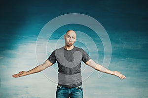 Young bald man in a gray T-shirt on a blue background spreads his arms