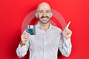 Young bald man holding floppy disk smiling happy pointing with hand and finger to the side