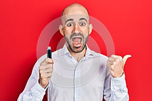 Young bald man holding removable memory usb pointing thumb up to the side smiling happy with open mouth