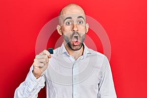 Young bald man holding sdxc card scared and amazed with open mouth for surprise, disbelief face