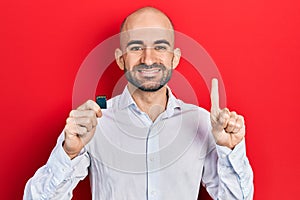 Young bald man holding sdxc card smiling with an idea or question pointing finger with happy face, number one