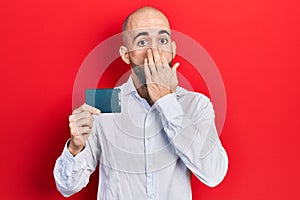 Young bald man holding ssd memory covering mouth with hand, shocked and afraid for mistake