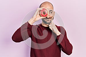 Young bald man holding tasty colorful doughnut on eye serious face thinking about question with hand on chin, thoughtful about