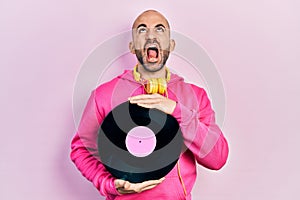 Young bald man holding vinyl disc angry and mad screaming frustrated and furious, shouting with anger looking up