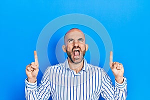 Young bald man pointing up with fingers angry and mad screaming frustrated and furious, shouting with anger looking up