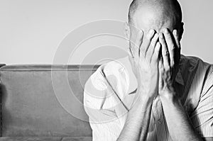 Young bald man in the shirt feeling depressed and miserable cover his face with his hands and cry in his room black and white.