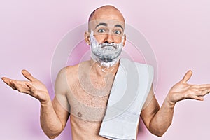 Young bald man shirtless shaving beard with foam clueless and confused expression with arms and hands raised