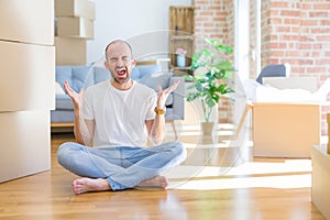 Young bald man sitting on the floor around cardboard boxes moving to a new home celebrating mad and crazy for success with arms