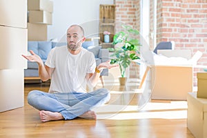 Young bald man sitting on the floor around cardboard boxes moving to a new home clueless and confused expression with arms and