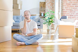 Young bald man sitting on the floor around cardboard boxes moving to a new home with hand on chin thinking about question, pensive