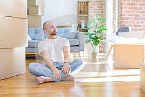 Young bald man sitting on the floor around cardboard boxes moving to a new home looking away to side with smile on face, natural