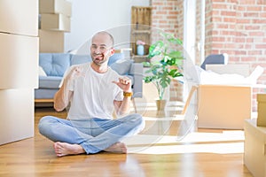 Young bald man sitting on the floor around cardboard boxes moving to a new home looking confident with smile on face, pointing