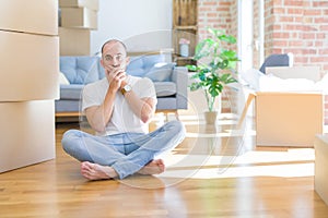 Young bald man sitting on the floor around cardboard boxes moving to a new home shocked covering mouth with hands for mistake