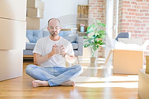 Young bald man sitting on the floor around cardboard boxes moving to a new home smiling with hands on chest with closed eyes and