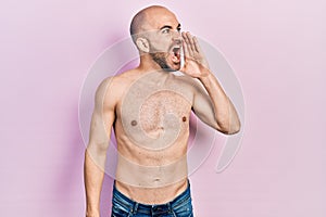 Young bald man standing shirtless shouting and screaming loud to side with hand on mouth