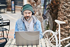 Young bald man using laptop and headphones sitting on table at coffee shop terrace