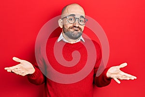 Young bald man wearing casual clothes and glasses clueless and confused expression with arms and hands raised