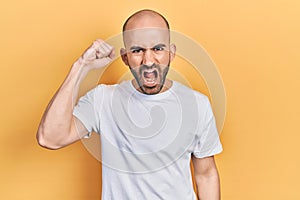 Young bald man wearing casual white t shirt angry and mad raising fist frustrated and furious while shouting with anger