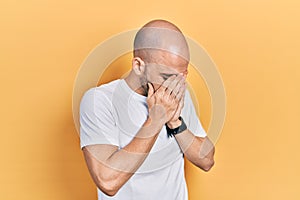 Young bald man wearing casual white t shirt with sad expression covering face with hands while crying