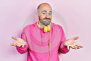 Young bald man wearing gym clothes and using headphones clueless and confused expression with arms and hands raised