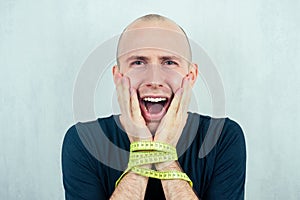 A young balding bald man yells and screams holding his head with the bound measuring tape tied hands. concept of diet