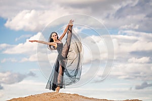 young ballerina in a black long dress stands in a graceful pose against a background of blue sky and clouds.