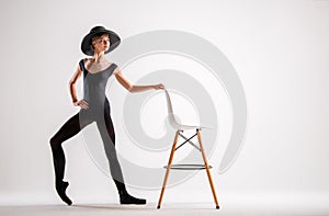 Young ballerina in black pointe shoes and an elegant hat flies along with chair on gray background