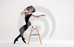 Young ballerina in black pointe shoes and elegant hat posing sitting on chair isolated on white background