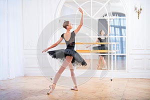 Young ballerina in a black tutu stands in graceful pose on pointe shoes in a large bright hall in front of a mirror