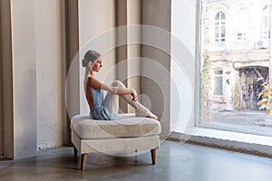 Young ballerina in blue dress sits by window in serene pose, reflecting quietly before ballet class
