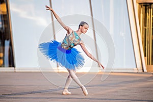 Young ballerina in a bright blue tutu is dancing against backdrop of cityscape