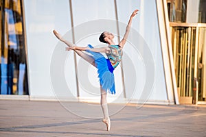 Young ballerina in a bright blue tutu is dancing against backdrop of cityscape