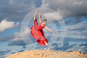 Young ballerina in a bright red long dress soars in a jump above the ground