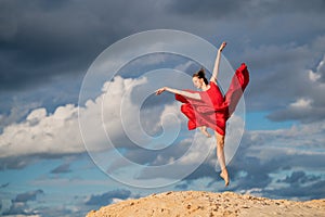 Young ballerina in a bright red long dress soars in a jump above the ground