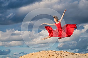 Young ballerina in a bright red long dress soars in a jump above the ground