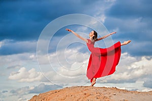 Young ballerina in a bright red long dress soars in a jump above the ground