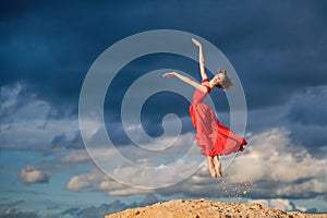 Young ballerina in a bright red long dress soars in a jump above the ground