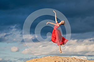 Young ballerina in a bright red long dress soars in a jump above the ground