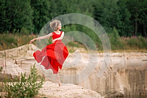 Young ballerina in a bright red long dress soars in a jump above the ground