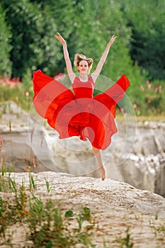 young ballerina in a bright red long dress soars in a jump above the ground