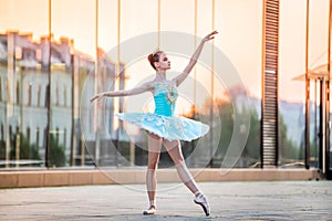 Young ballerina is dancing on pointe shoes in delicate mint blue tutu against the backdrop of reflection of sunset in the city