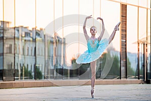 Young ballerina is dancing on pointe shoes in delicate mint blue tutu against the backdrop of reflection of sunset in the city