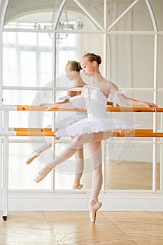 Young ballerina is doing classical ballet at a ballet barn in beautiful white room in front of a mirror