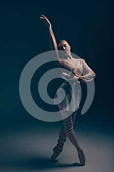 Young ballerina in dress and pointe shoes dancing against blue background