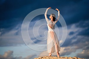 Young ballerina in a light long white dress stands in an arabesque
