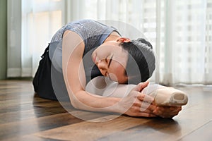 Young ballerina in pointe shoes is sitting on the floor, warming up before dance class practicing ballet in dance school