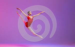 A young ballerina in a red leotard performs a split jump on a lilac background